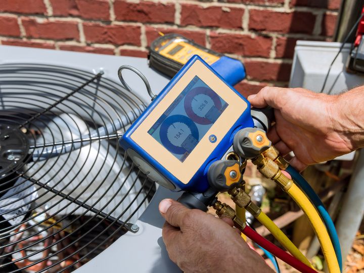A person holds a digital HVAC manifold gauge, connected with colored hoses to an outdoor air conditioning unit, checking pressure readings. A brick wall and another tool are visible in the background.