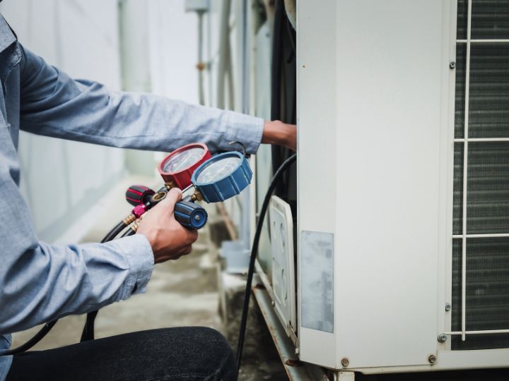 A technician in a blue shirt uses a pressure gauge to check or service an outdoor air conditioning unit. The technicians face is not visible, and the focus is on their hands and the equipment.