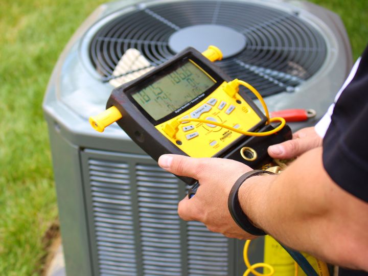 A person holds a digital manifold gauge while checking an outdoor air conditioning unit, measuring and monitoring the system’s performance and refrigerant levels.