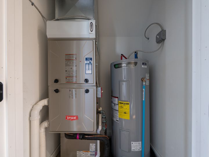 A home utility closet containing a gray Bryant furnace and a tall AO Smith water heater, both installed side by side with visible pipes and wiring against white walls.