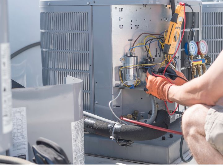 A technician wearing orange gloves uses tools to repair or inspect the wiring and components inside an outdoor air conditioning unit. Electrical meters and gauges are attached for diagnostics.