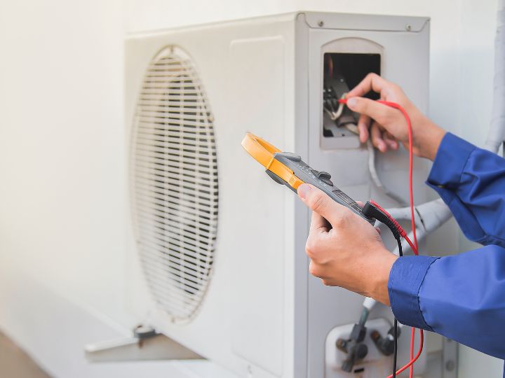 A technician in a blue uniform is using a digital multimeter to check or repair an outdoor air conditioning unit, holding the probes near the unit’s control panel.