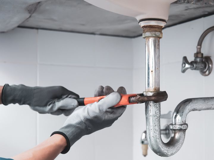 A person wearing gray gloves uses a wrench to tighten or loosen a metal pipe under a sink in a bathroom or kitchen.